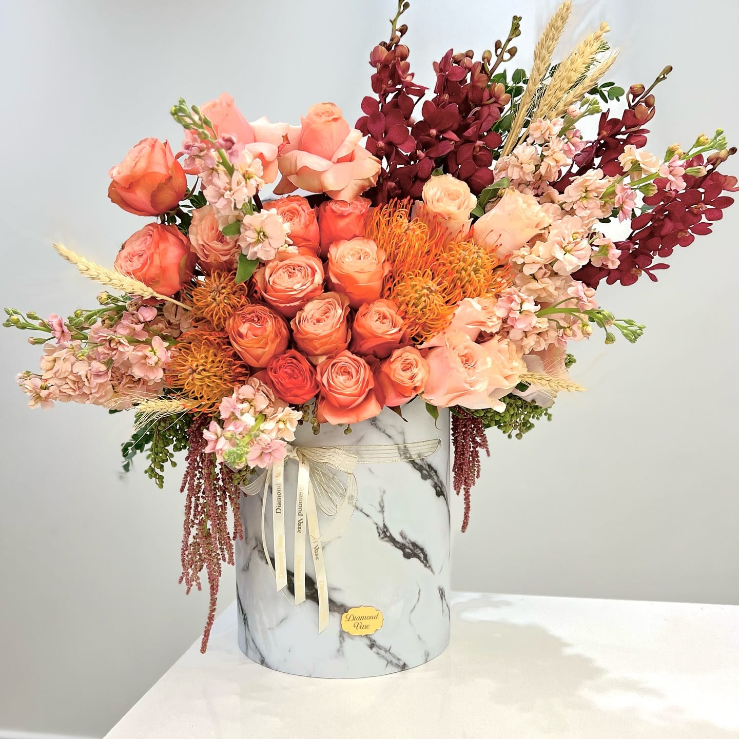Coral and peach garden roses, orange protea, burgundy orchids and wheat in a marble-print Diamond Vase box