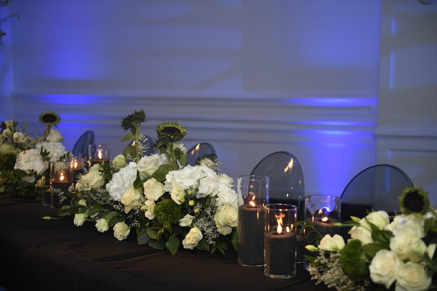 Low runner of white roses and hydrangea with black sand candles burning in glass cylinders
